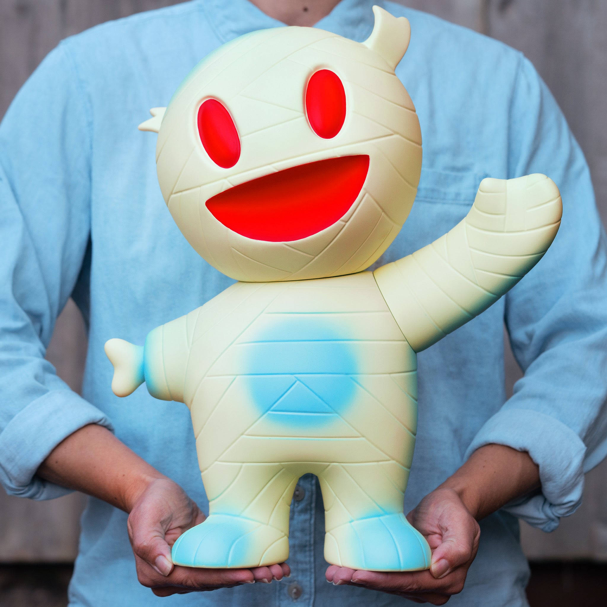 Person in a blue shirt holds a large glow in the dark mummy boy with red eyes and a wide red mouth.