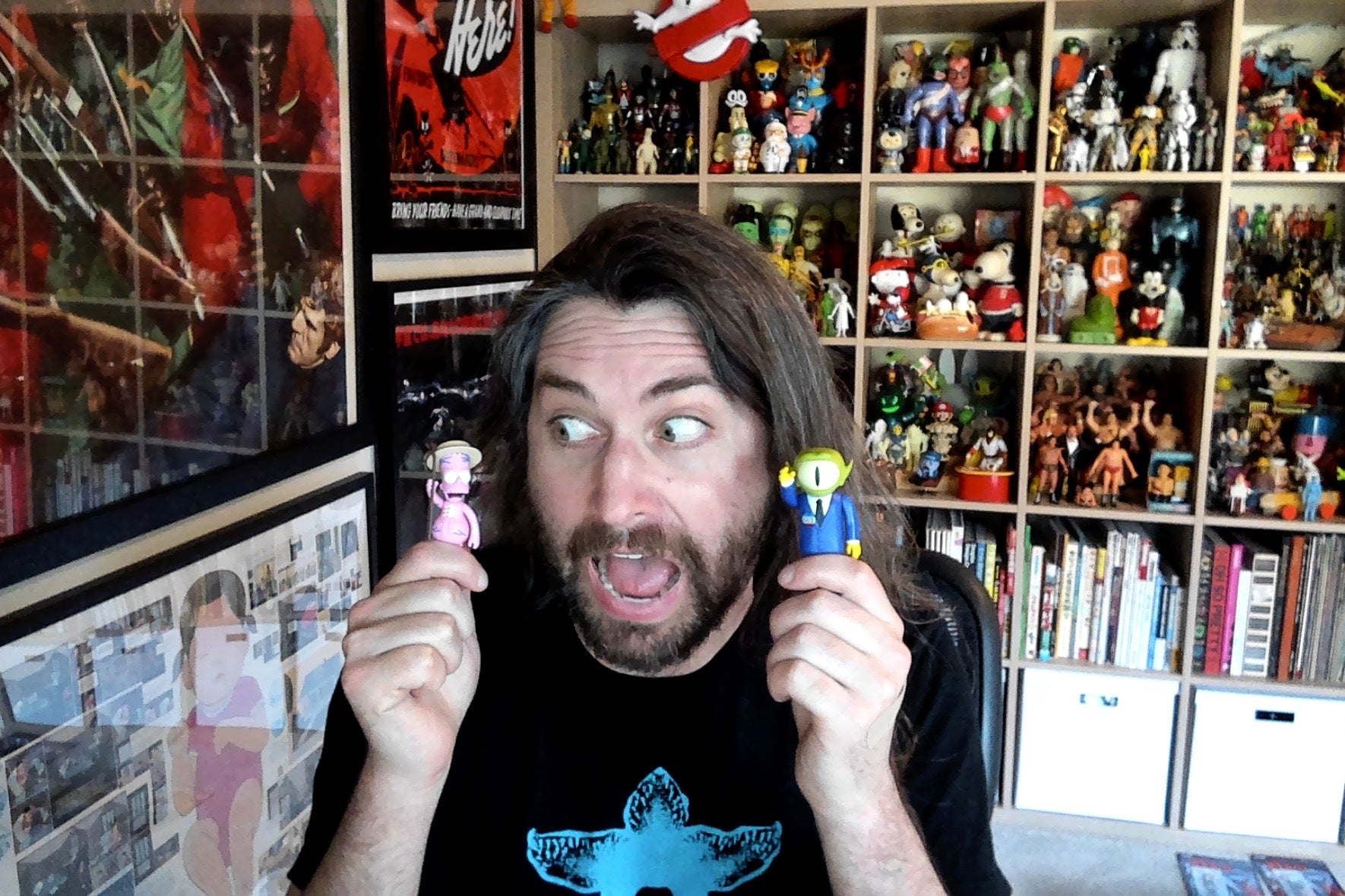 Man with long hair and beard holds two small collectible figures in a room full of toy shelves
