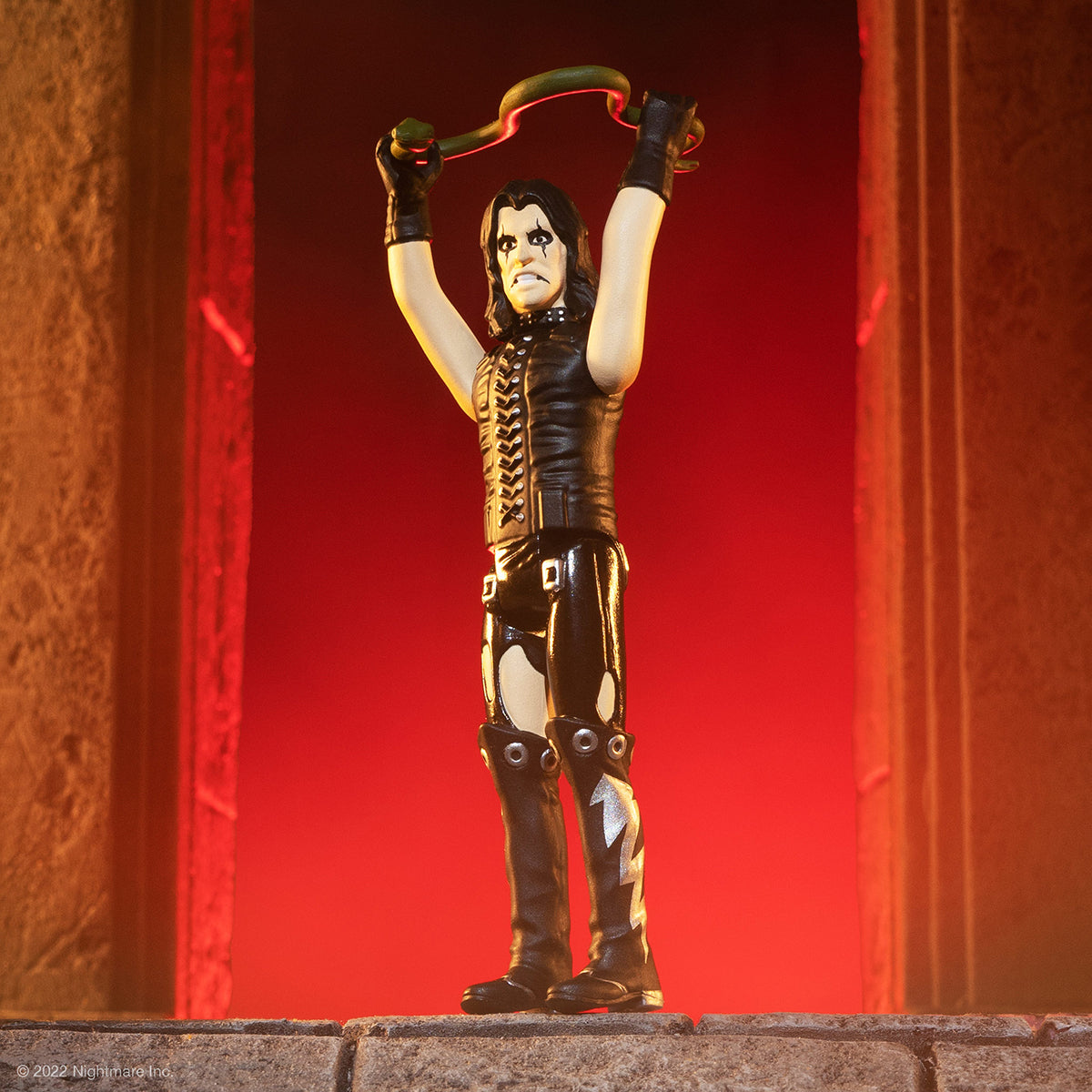 Rock icon Alice Cooper style action figure in black leather gear raises both arms gripping a green snake like prop on a dramatic red stage