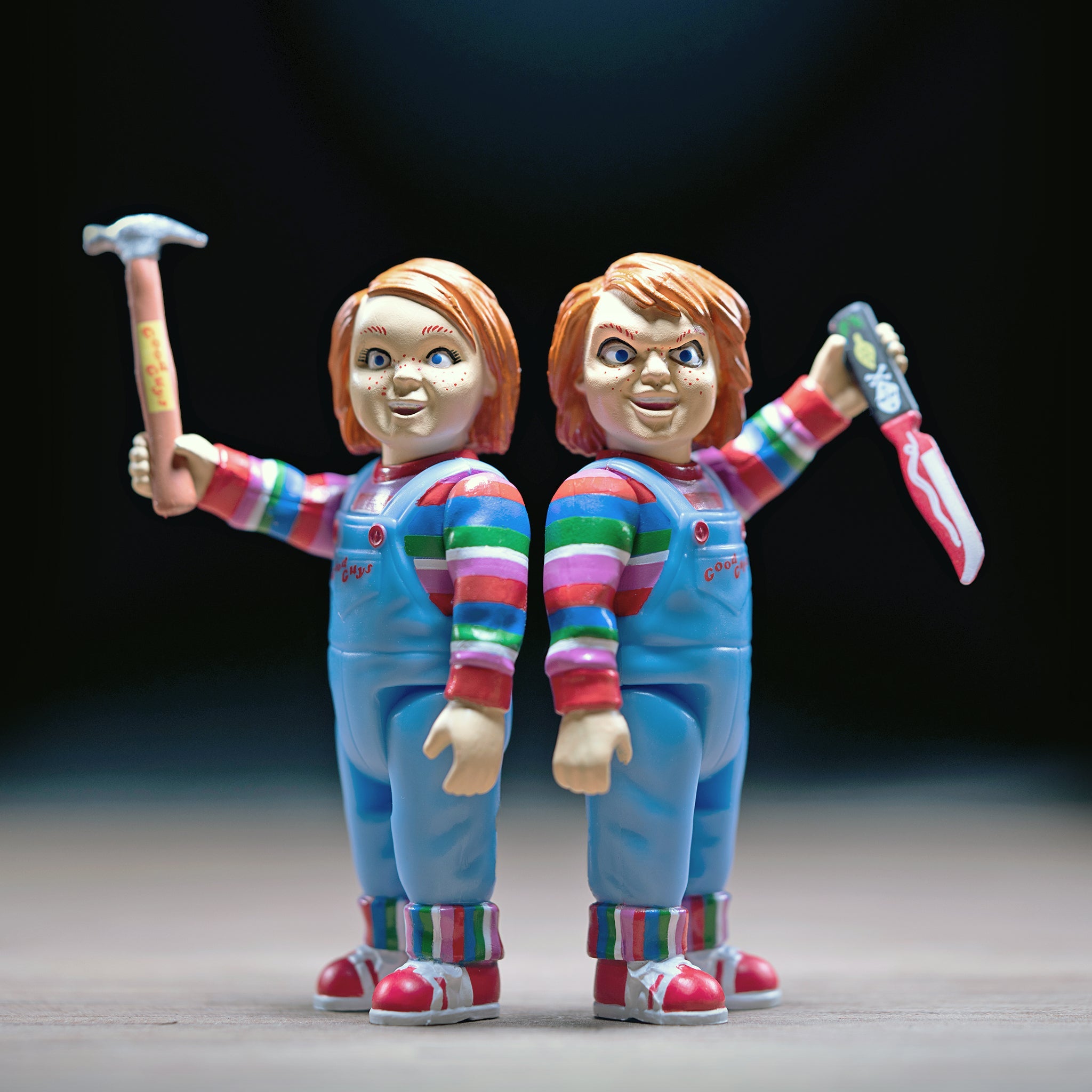 Two horror movie dolls with red hair and blue overalls posing against a dark background
