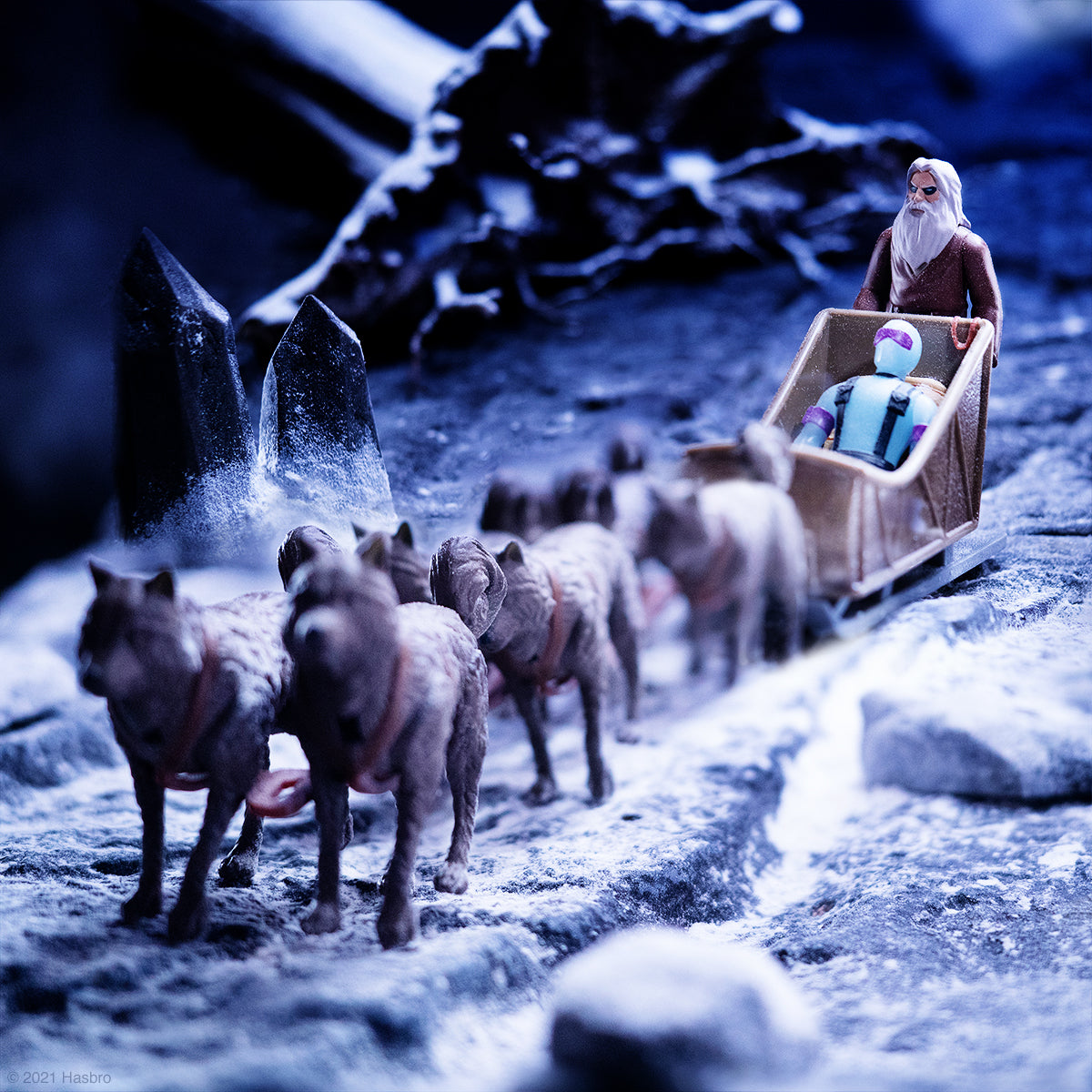 Camel caravan pulls a wooden wagon through a snowy rocky landscape with a bearded traveler at the reins.
