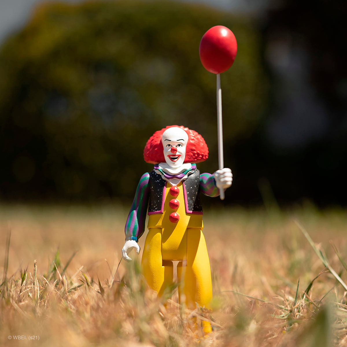 Clown figure in yellow pants and colorful shirt stands in a sunlit field holding a red balloon on a stick