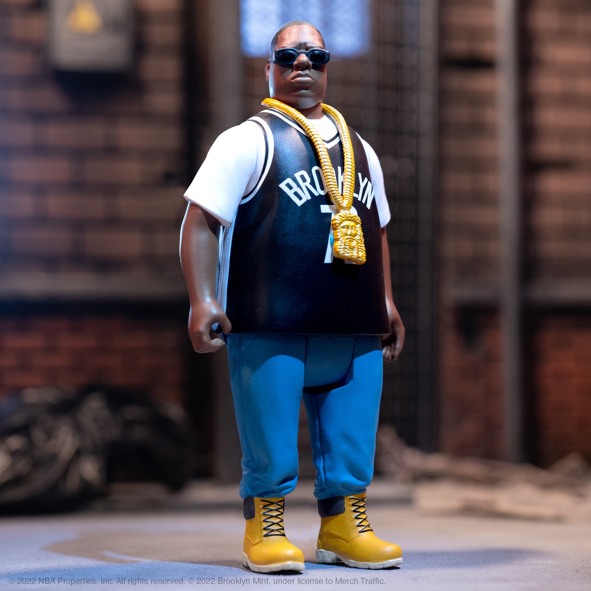 Notorious B.I.G. action figure wearing a Brooklyn jersey with a thick gold chain against a brick backdrop.