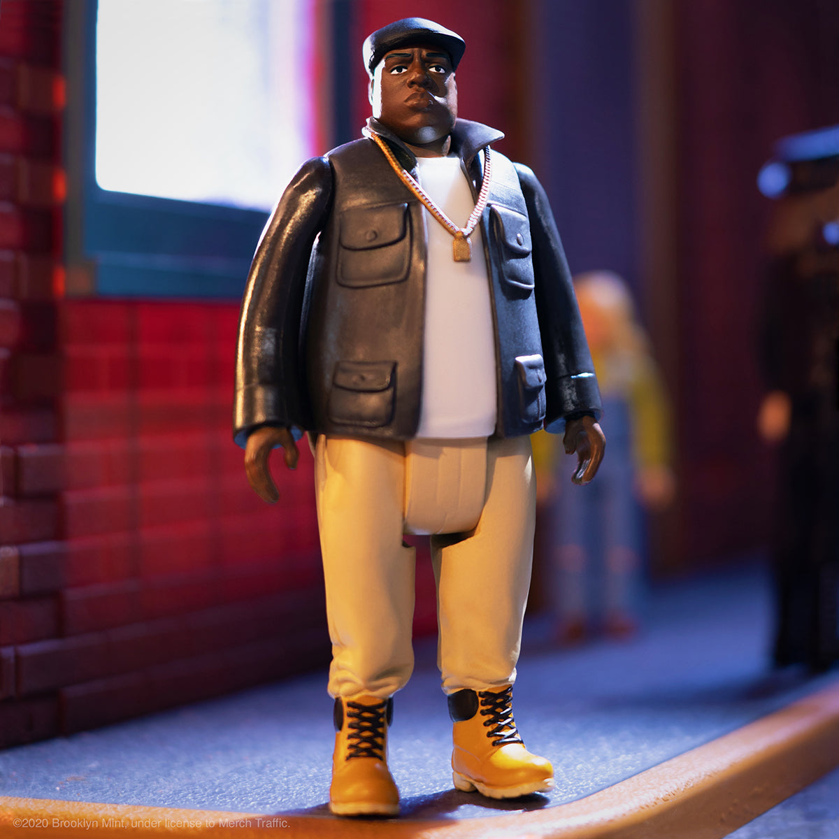 Notorious B I G action figure on a city sidewalk wearing a cap, white shirt, dark jacket and tan pants with a gold chain.