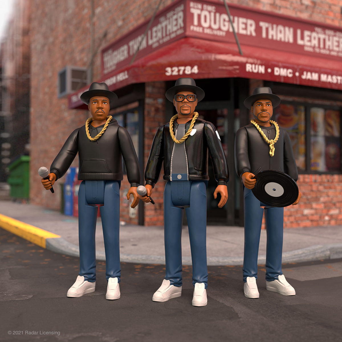 Three Run-DMC action figures in black outfits with gold chains stand on a city street in front of a brick storefront.