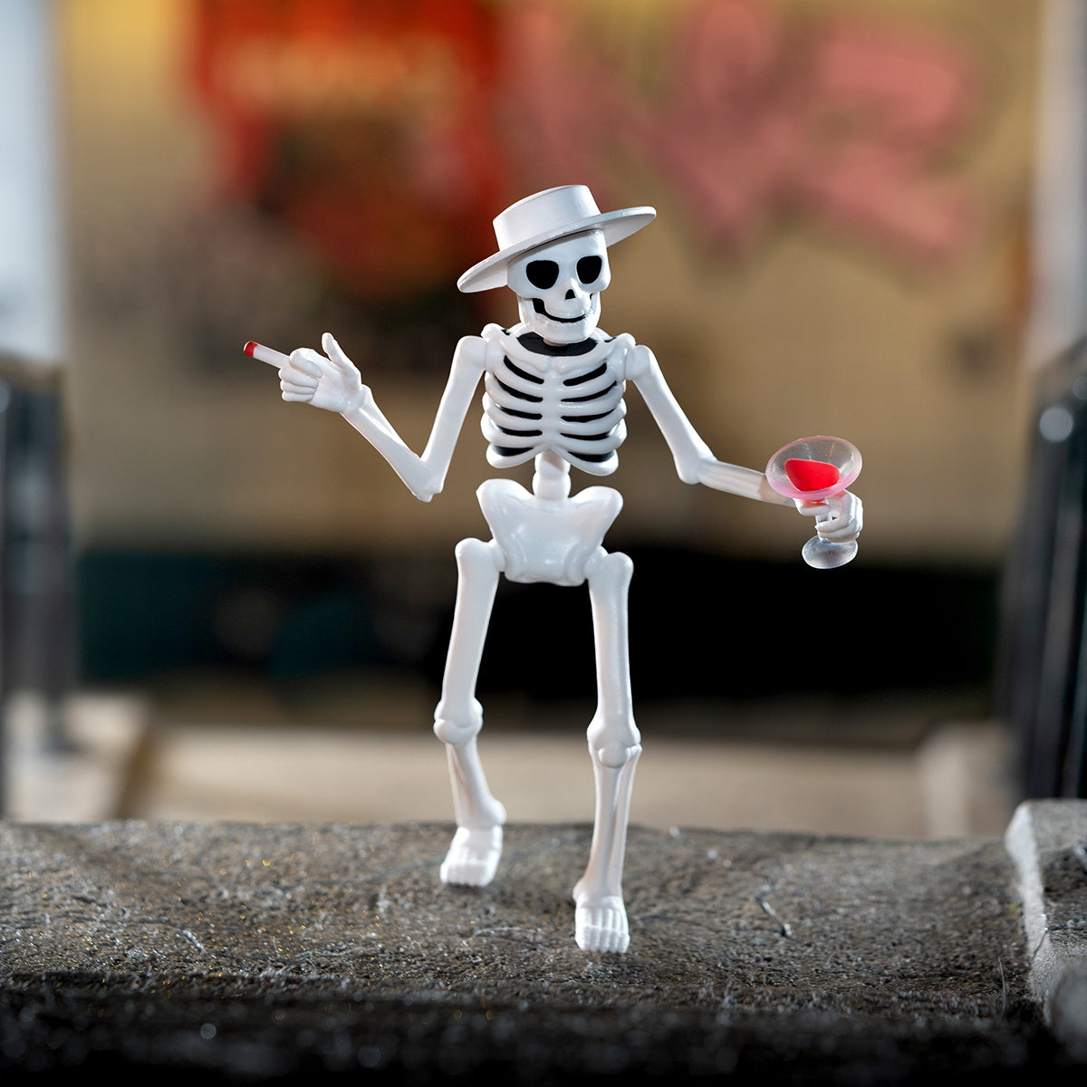 White skeleton action figure wearing a fedora holds a cigarette in one hand and a wine glass in the other.