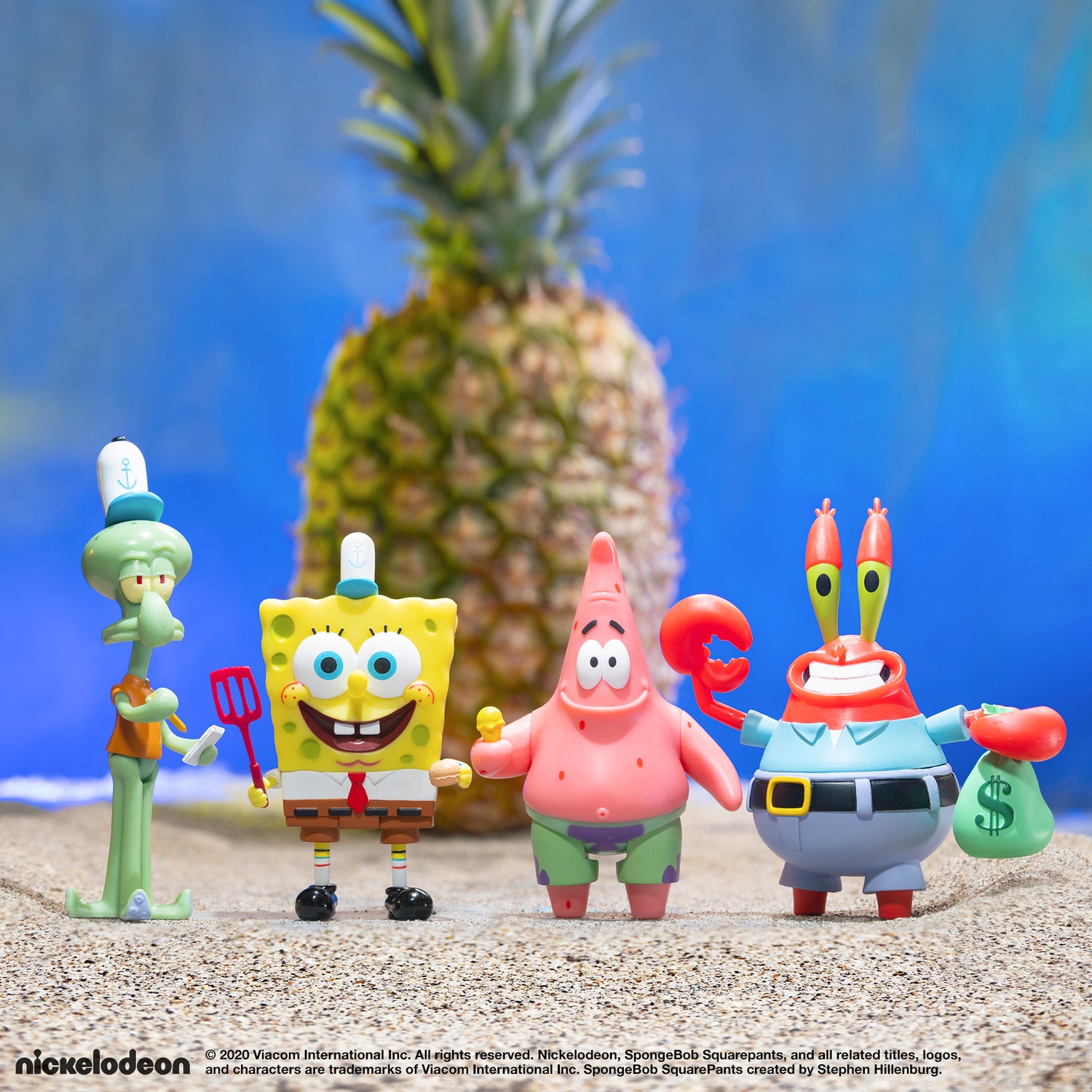 SpongeBob SquarePants toy figures posed on a sandy beach with a tall pineapple in the background