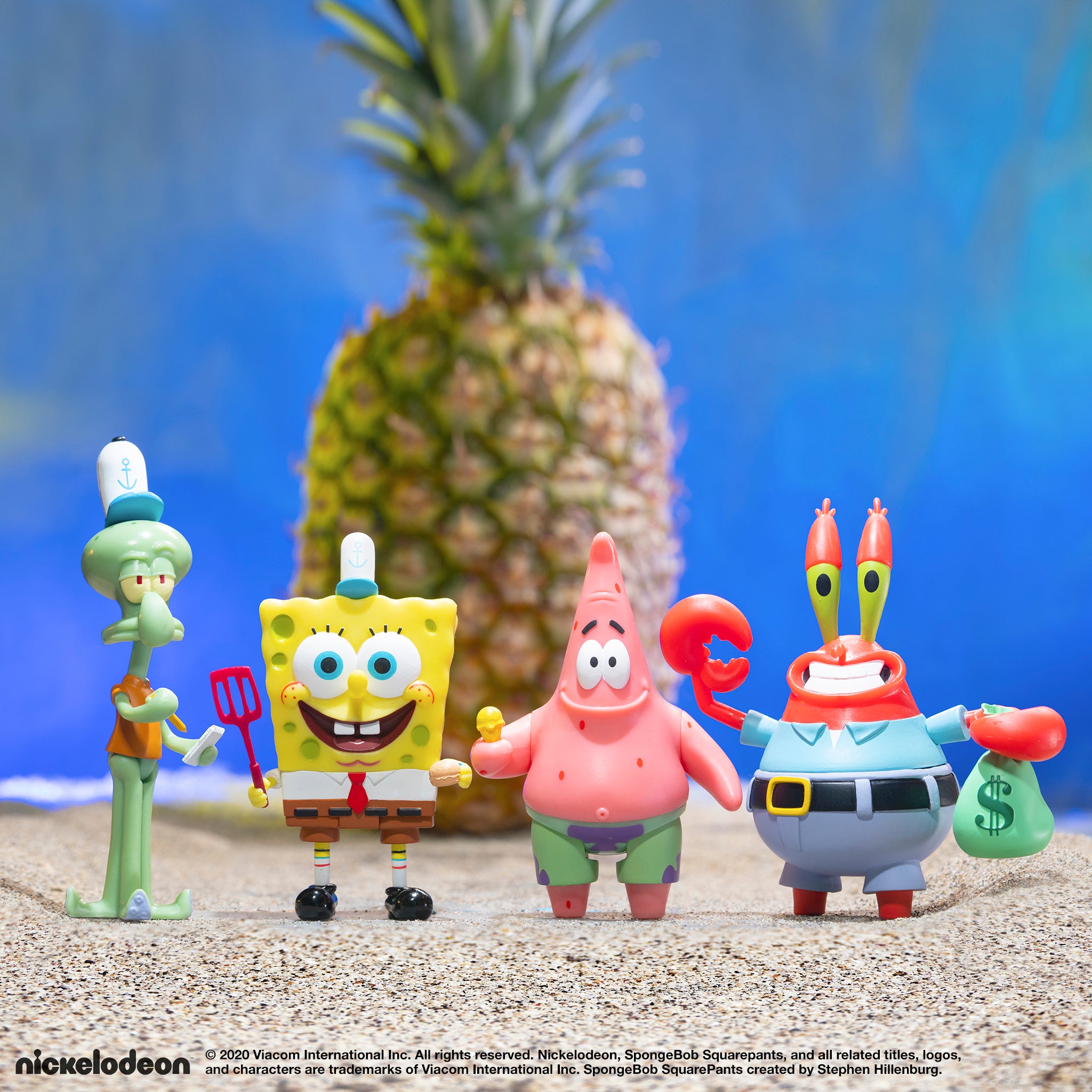 SpongeBob SquarePants toy figures posed on a sandy beach with a tall pineapple in the background