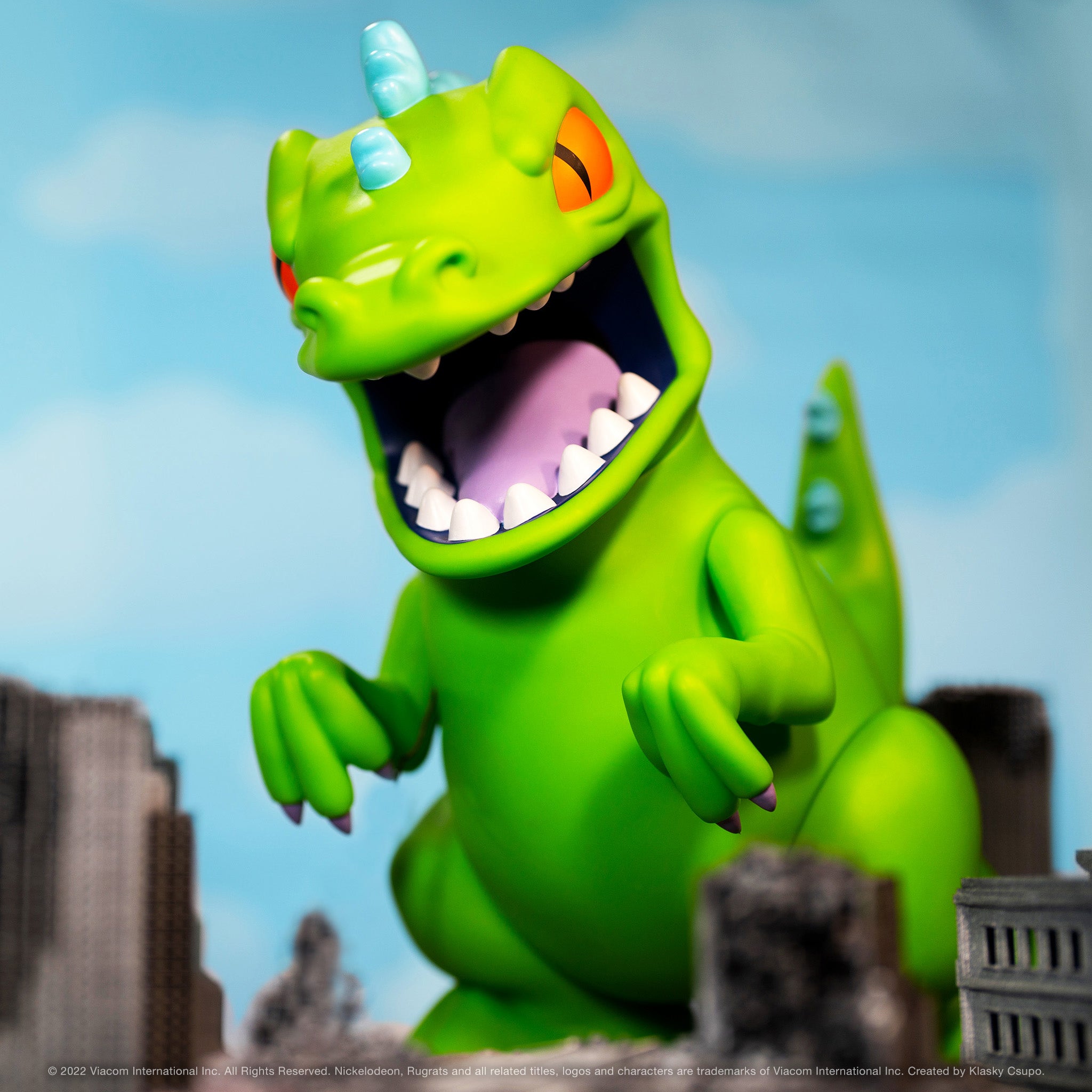 Gigantic green Reptar toy with an open mouth and sharp teeth against a blurred cityscape.