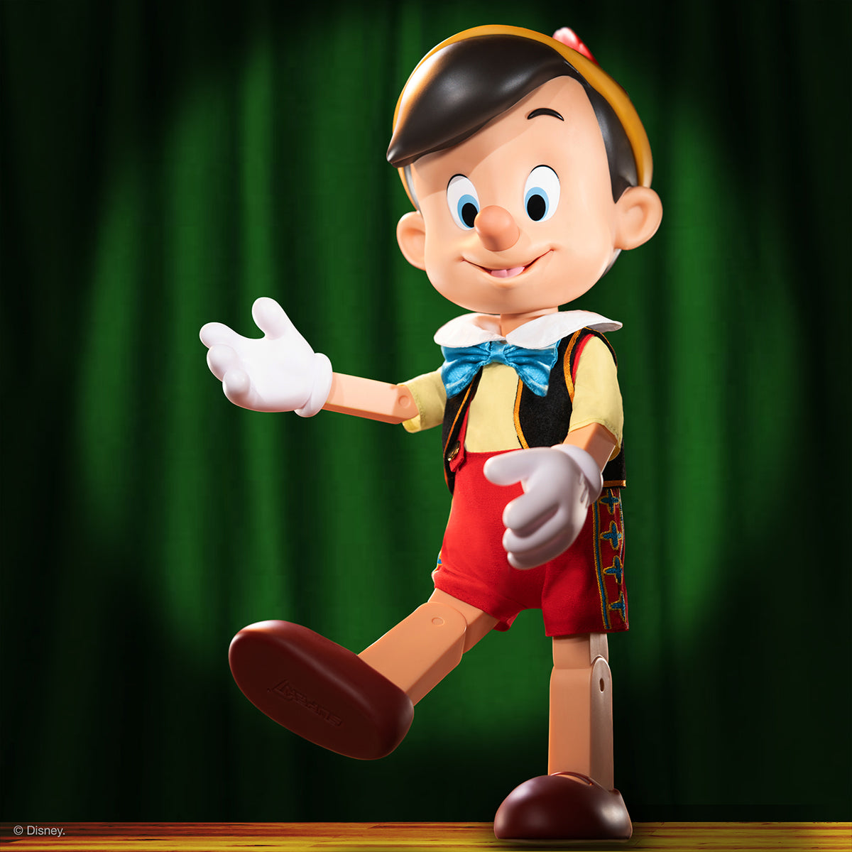 Disney Pinocchio supersize vinyl figure with yellow hat blue bow tie red shorts and outstretched arms against a green curtain.