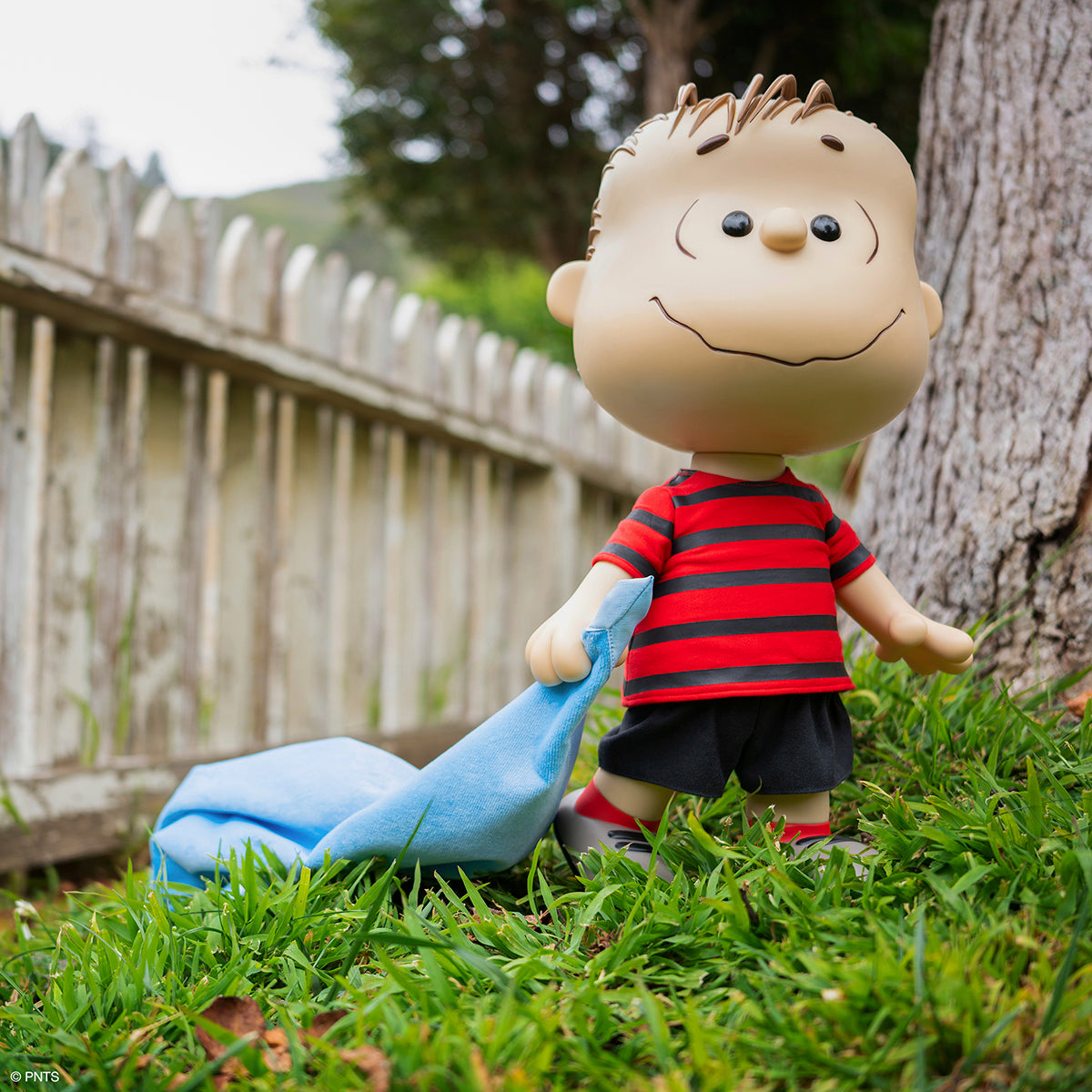 Supersize Linus toy outdoors in a red and black striped shirt holding a blue blanket beside a tree.