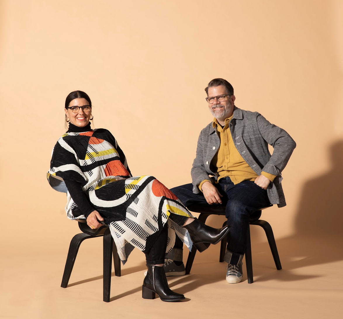 Two people sit on black chairs against a peach studio backdrop; the woman wears a colorful poncho, the man a mustard shirt and gray jacket.