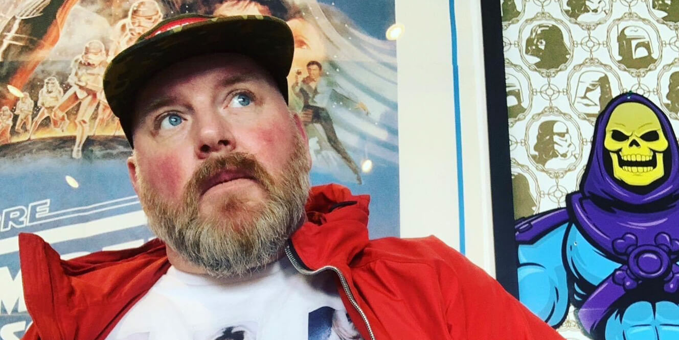 Portrait of Daniel Sant wearing a red jacket and camo cap, looking up with vintage posters in the background.