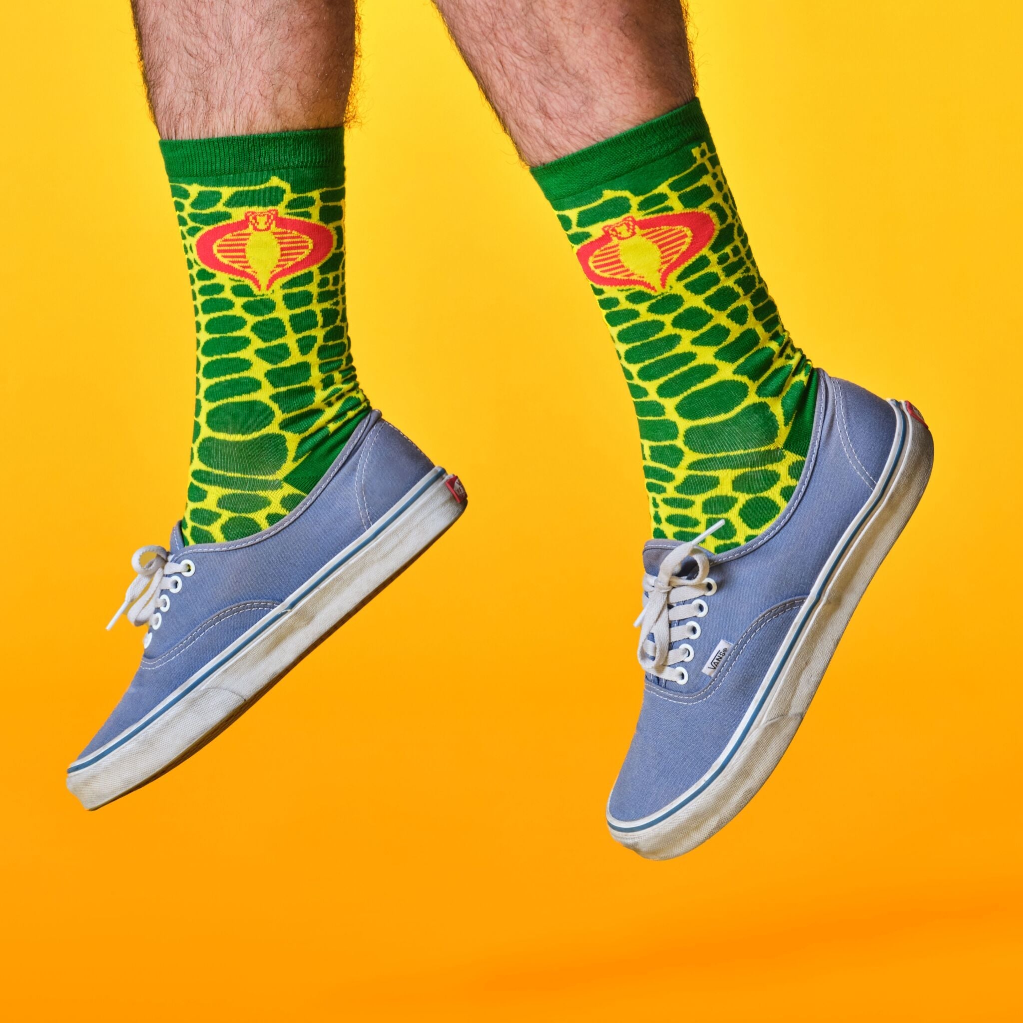 Legs wearing green snake print socks with crown motif and blue sneakers against an orange backdrop