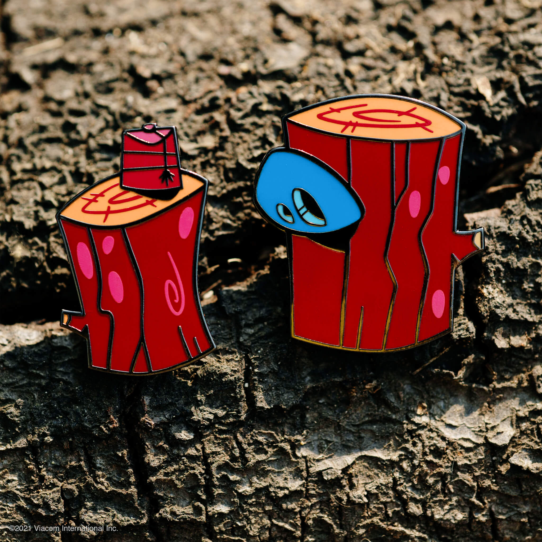 Close up of enamel pins on a dirt surface featuring red orange box shapes with a blue face.
