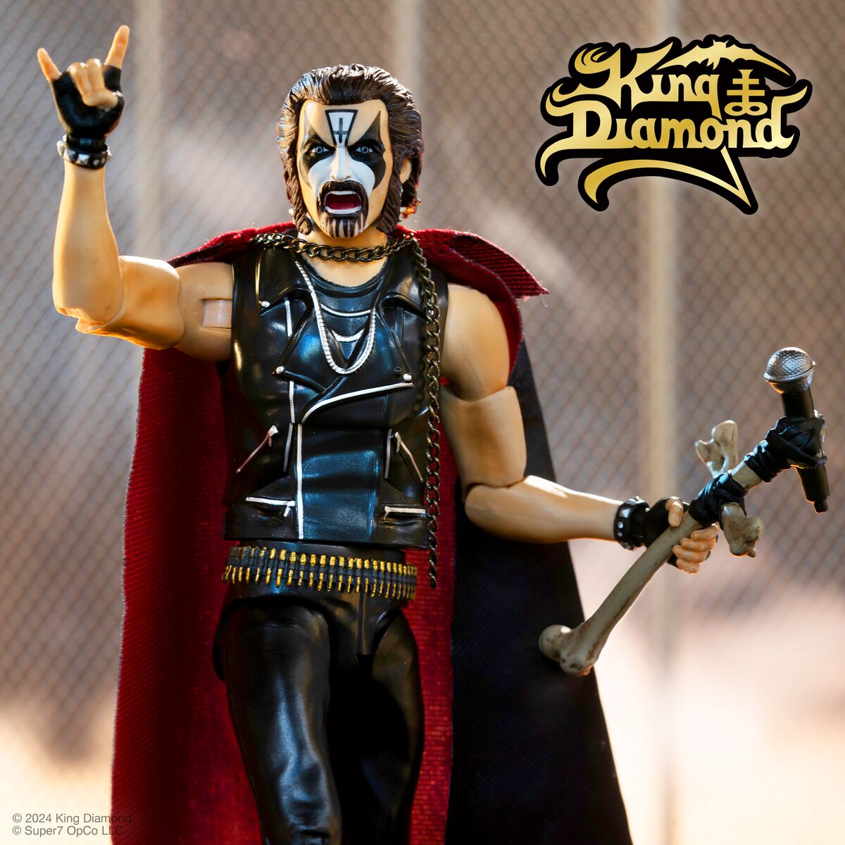 King Diamond style action figure in black leather outfit with cape holds a microphone on a stage backdrop.