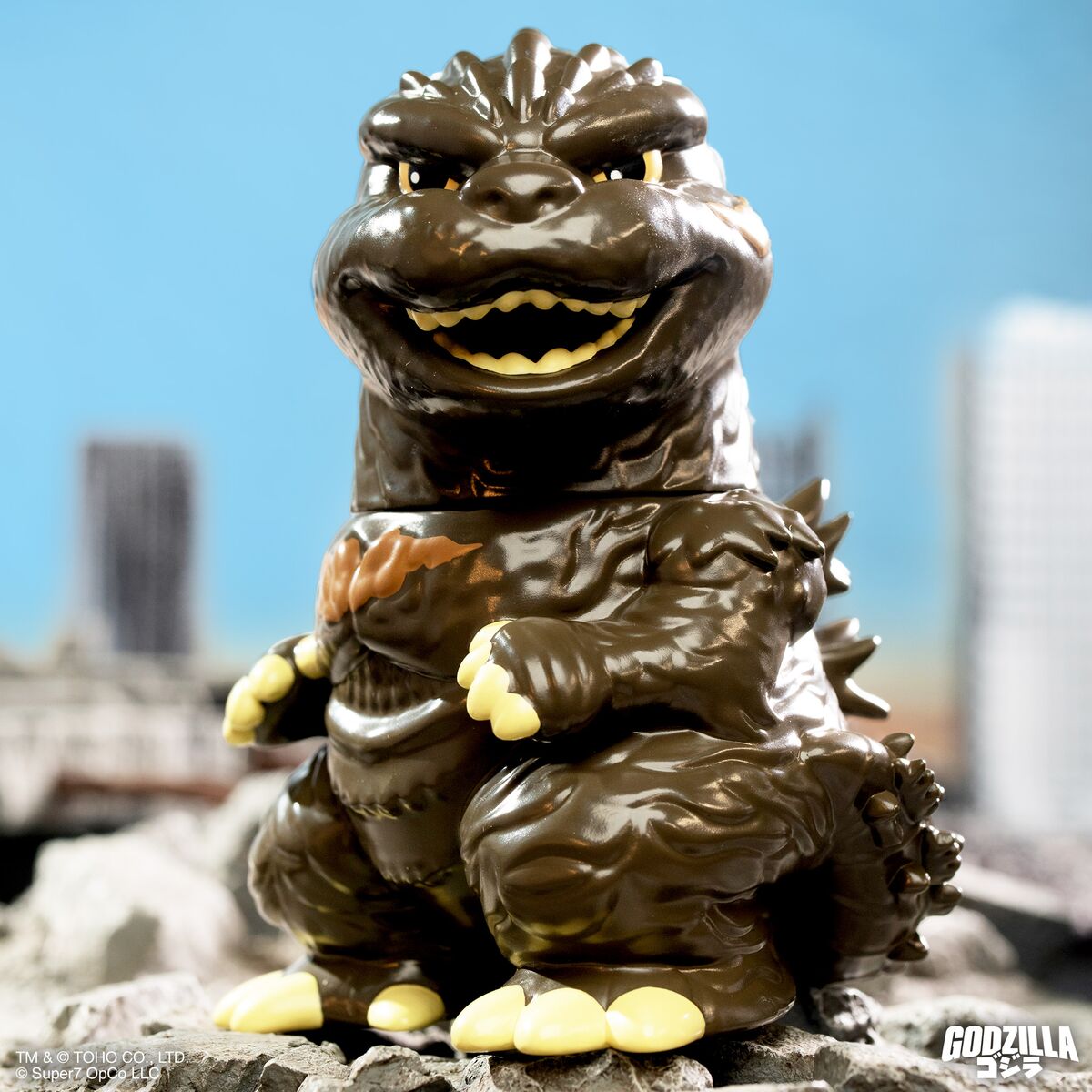Glossy brown Godzilla figure with bared teeth standing on rocky ground with a blue sky and city skyline behind