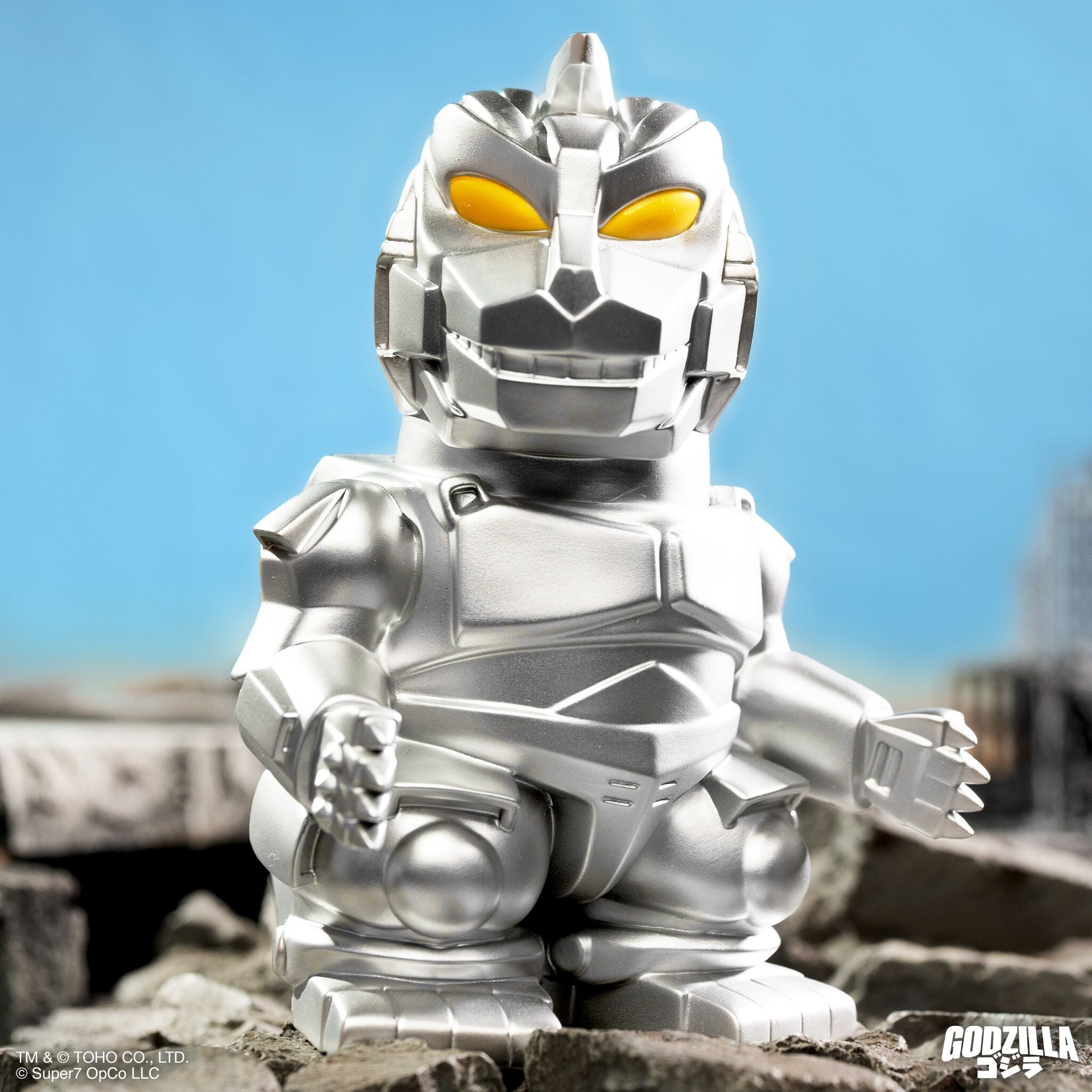 Close-up of a metallic silver Godzilla figure with yellow eyes standing on rocky ground against a bright blue sky.