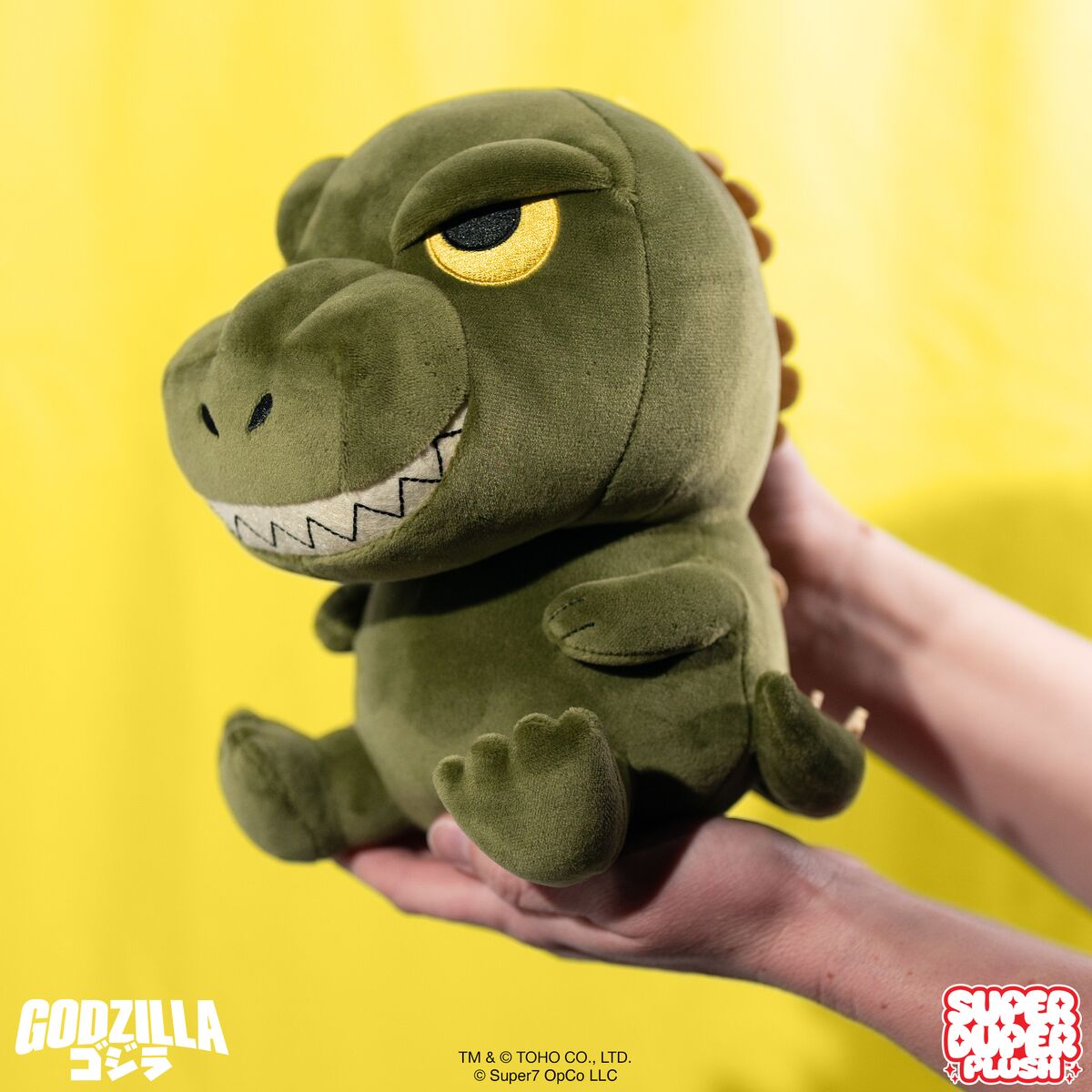 Green Godzilla plush held against a bright yellow backdrop