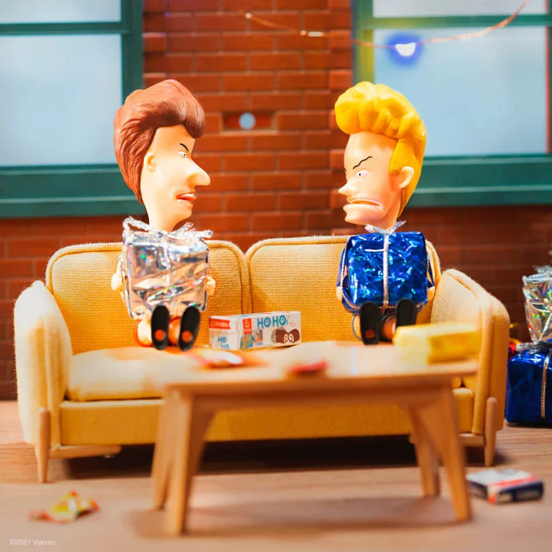Two ventriloquist dolls sit on a yellow sofa with a small table and wrapped gifts in a cozy living room setup.