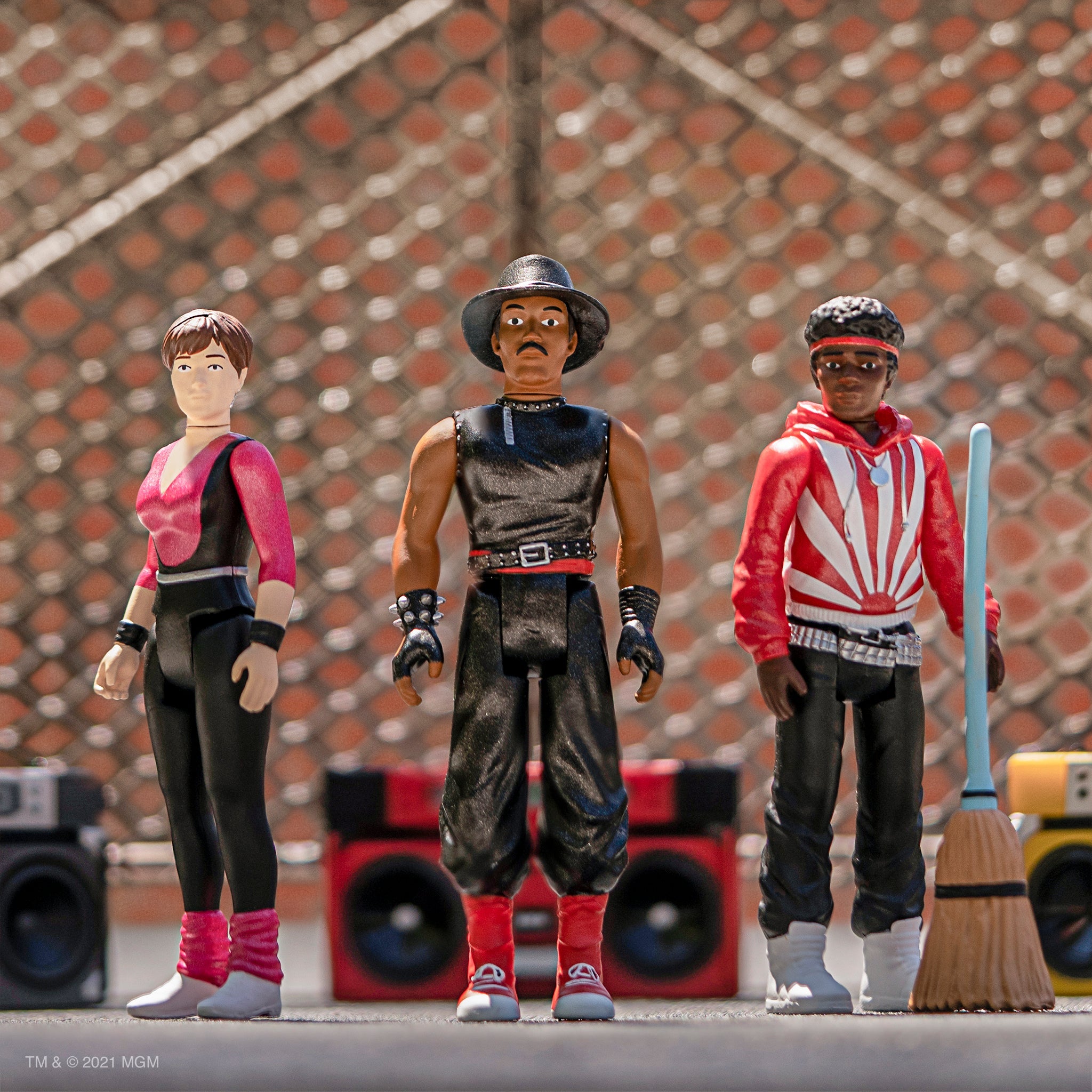 Three action figures from Breakin stand together in bold outfits against a blurred chain link background.