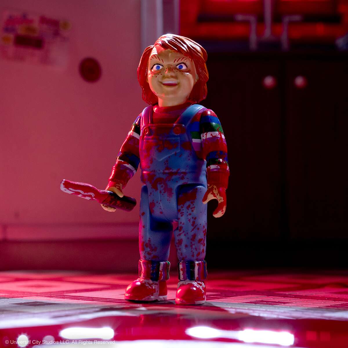 Blood-splattered Chucky doll in red overalls holding a plastic knife on a pink-lit stage