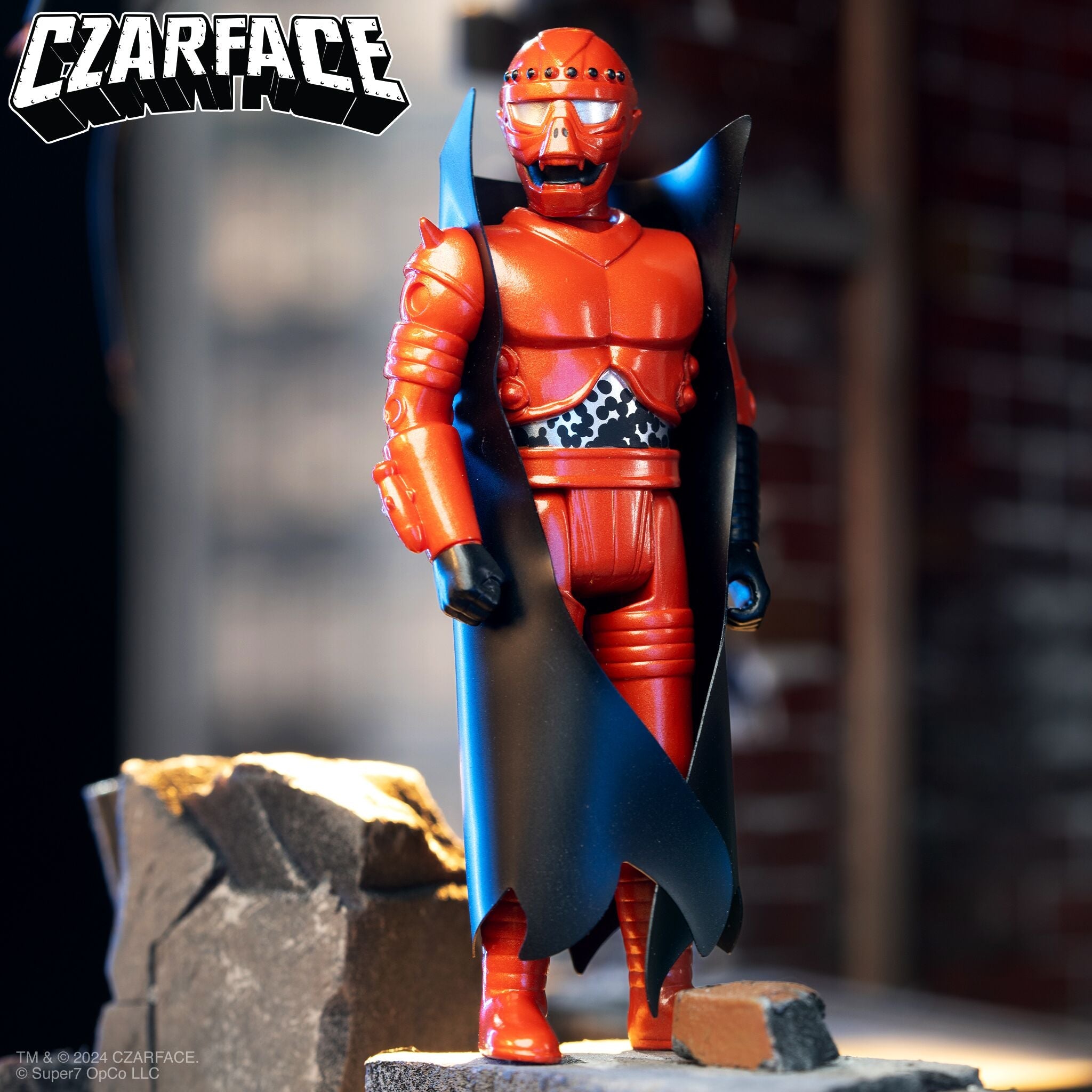 Red armored action figure with a black cape standing on a rocky pedestal