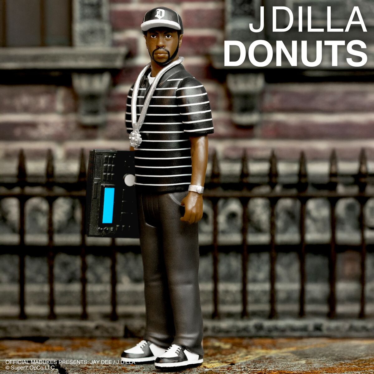 J Dilla action figure wearing a striped shirt and cap with a chunky chain, standing beside a boombox against an urban brick backdrop