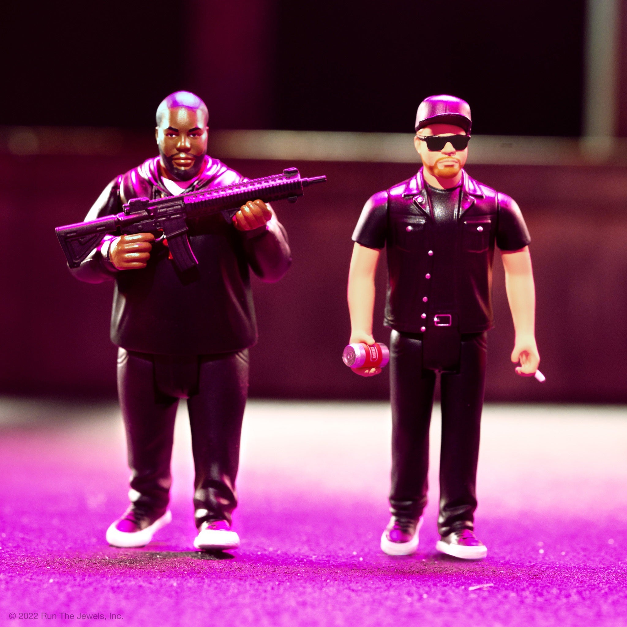 Two action figures in black outfits with one holding a rifle and the other wearing sunglasses against a purple backdrop