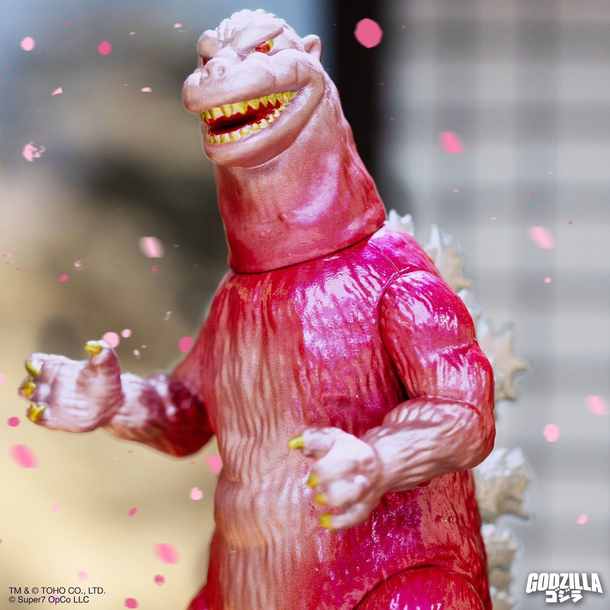 Close-up of a pink Godzilla figure with yellow teeth, set against a blurred background with pink confetti.