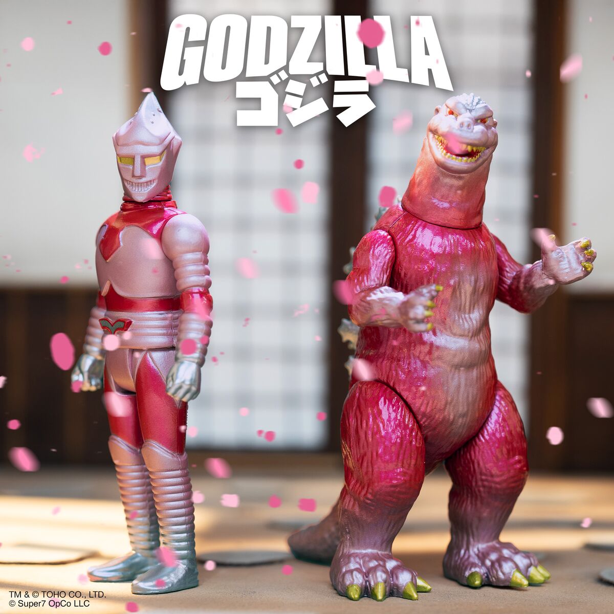 Two monster figures posed on a surface with pink confetti and the Godzilla logo in the background.