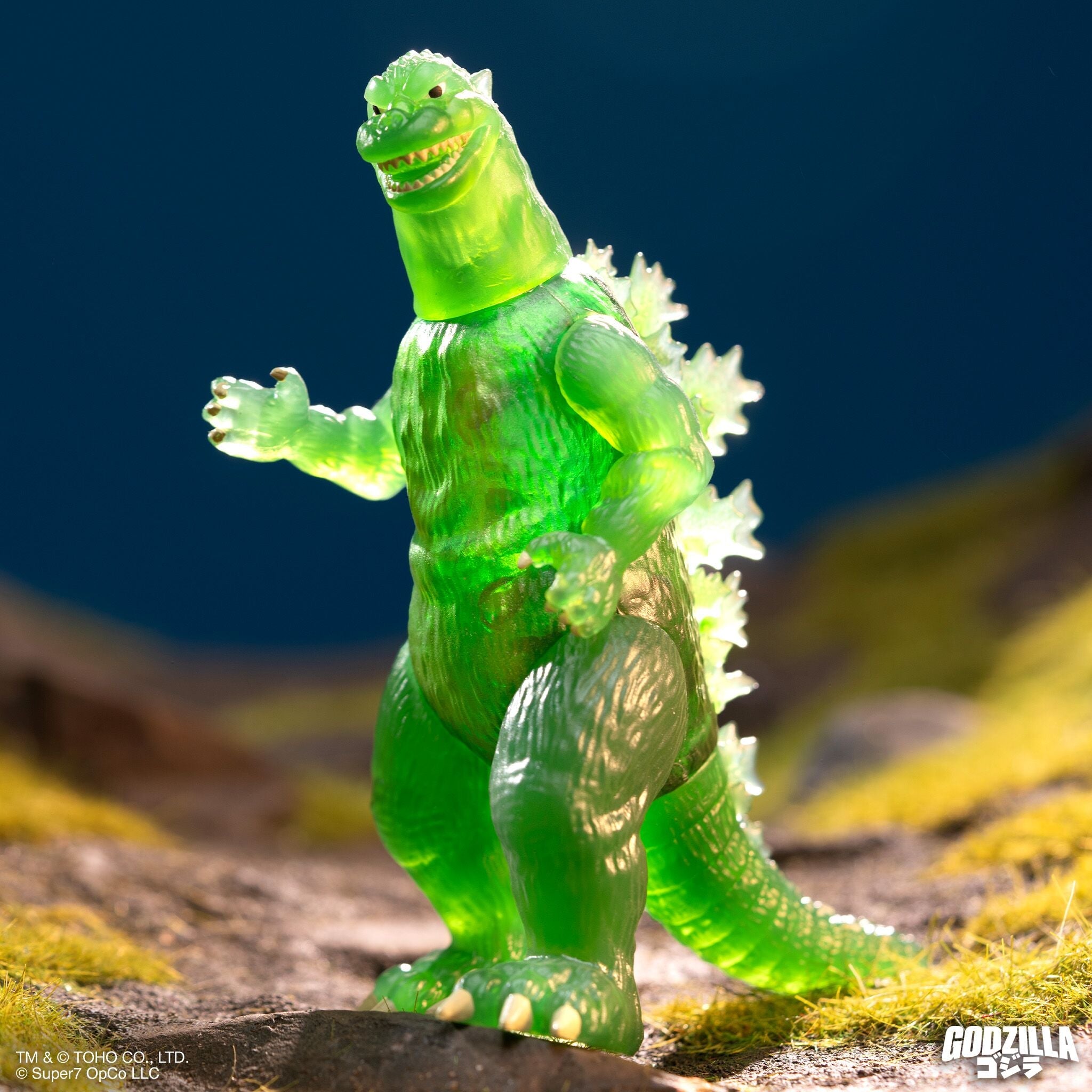Translucent green Godzilla figure with white belly and yellow dorsal spines on a rocky diorama