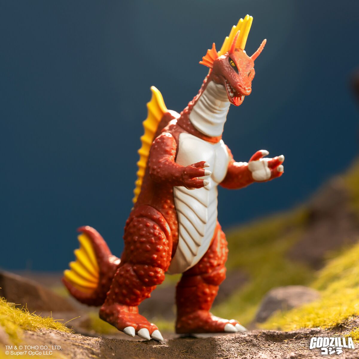 Red Godzilla figure with orange dorsal spines standing on a rocky diorama