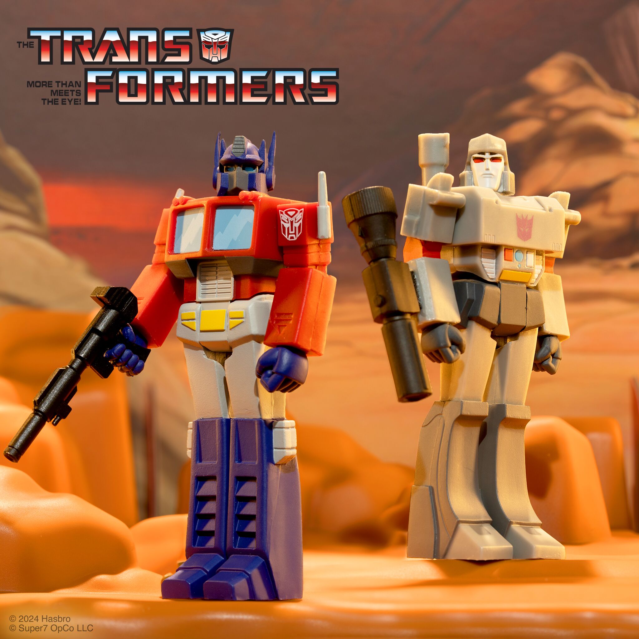 Two Transformer action figures stand in a desert setting with a bold Transformers logo in the corner
