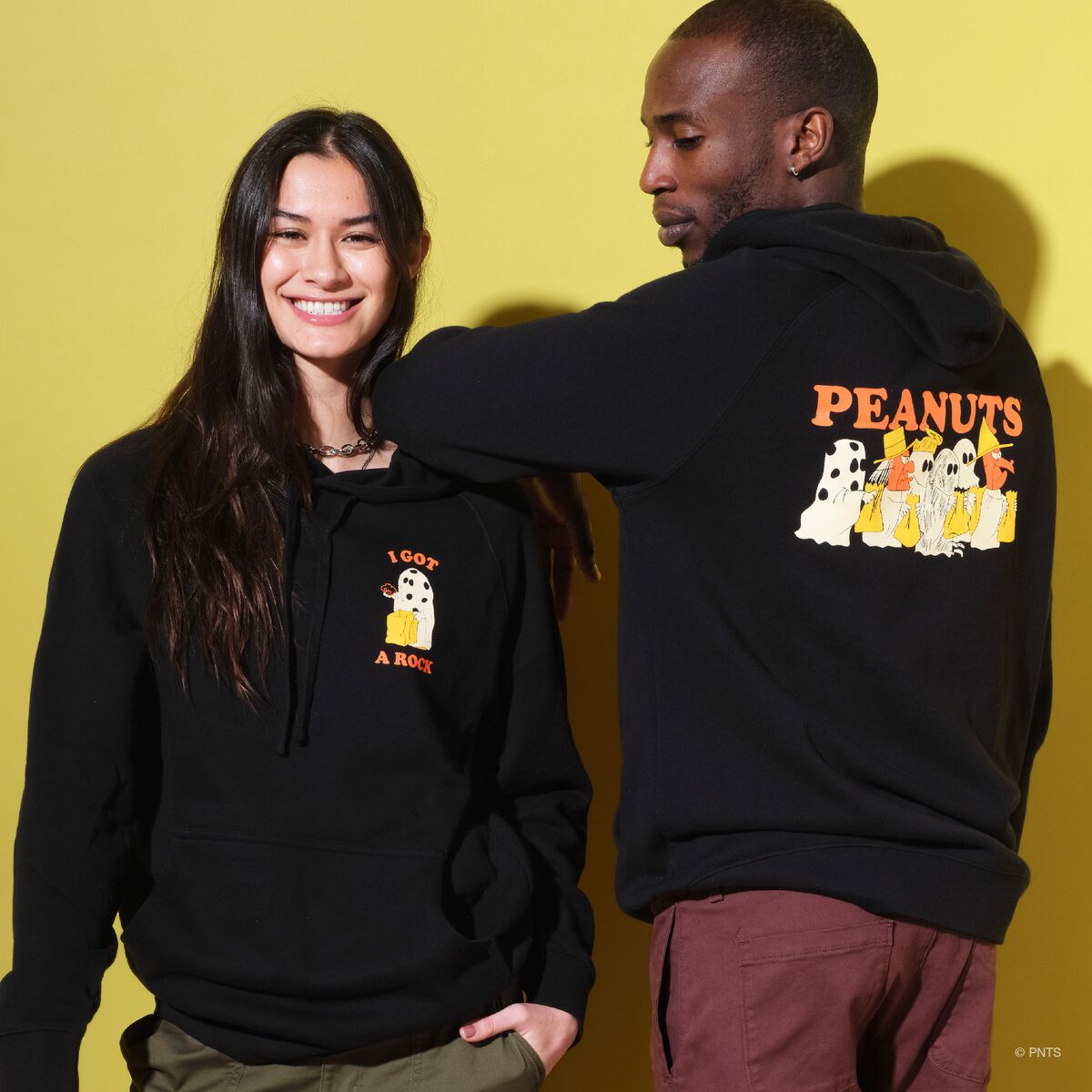 Two adults wear Peanuts themed hoodies posing against a bright yellow backdrop