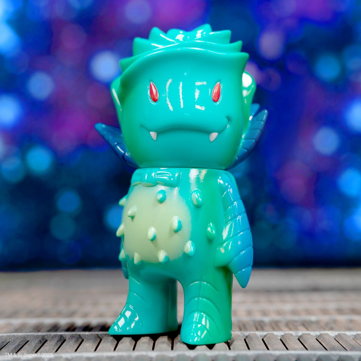 Turquoise dragonlike action figure with red eyes and white fangs, spiked back, standing on a wooden surface.