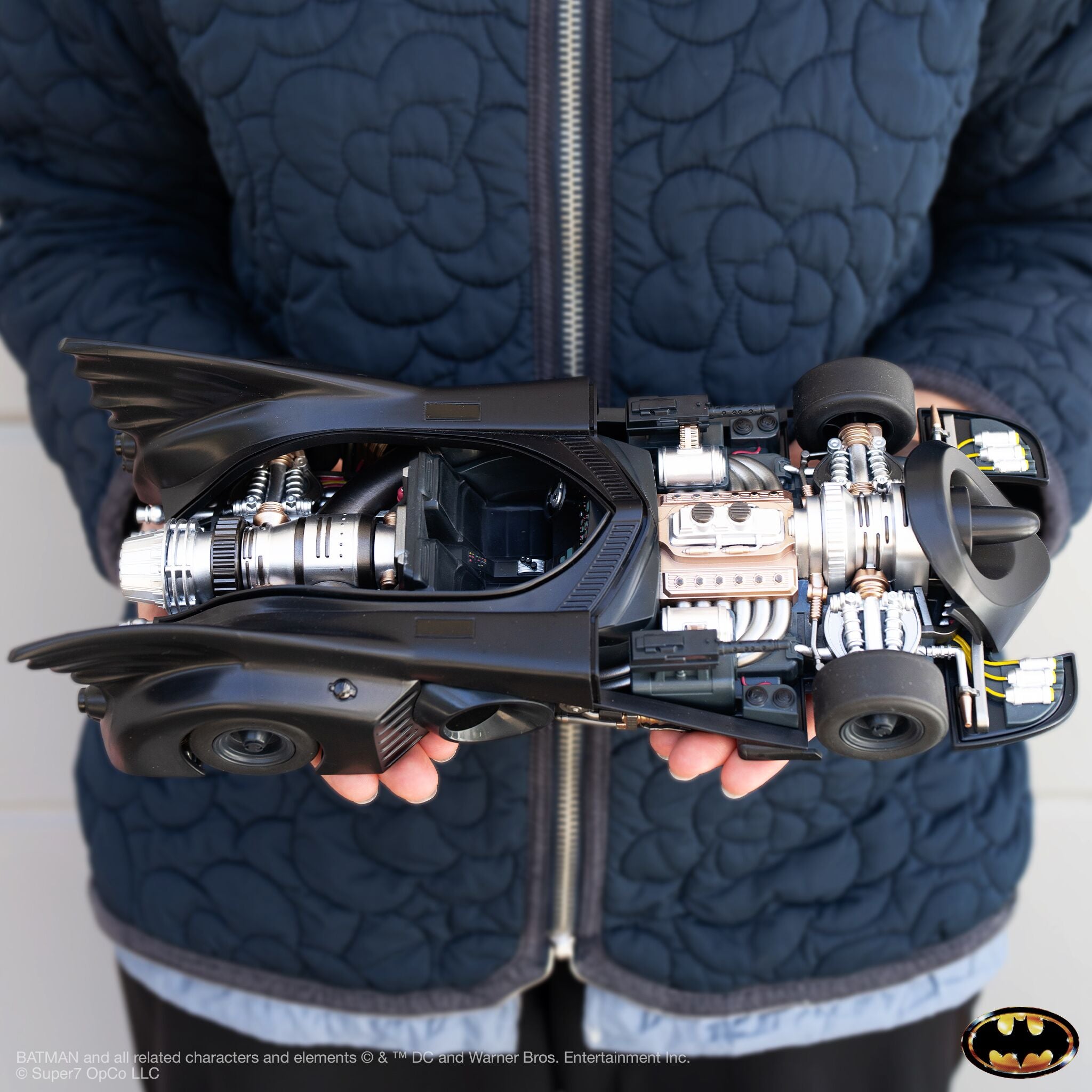 Person in a quilted navy jacket holds a Batmobile open to reveal its exposed engine and interior details