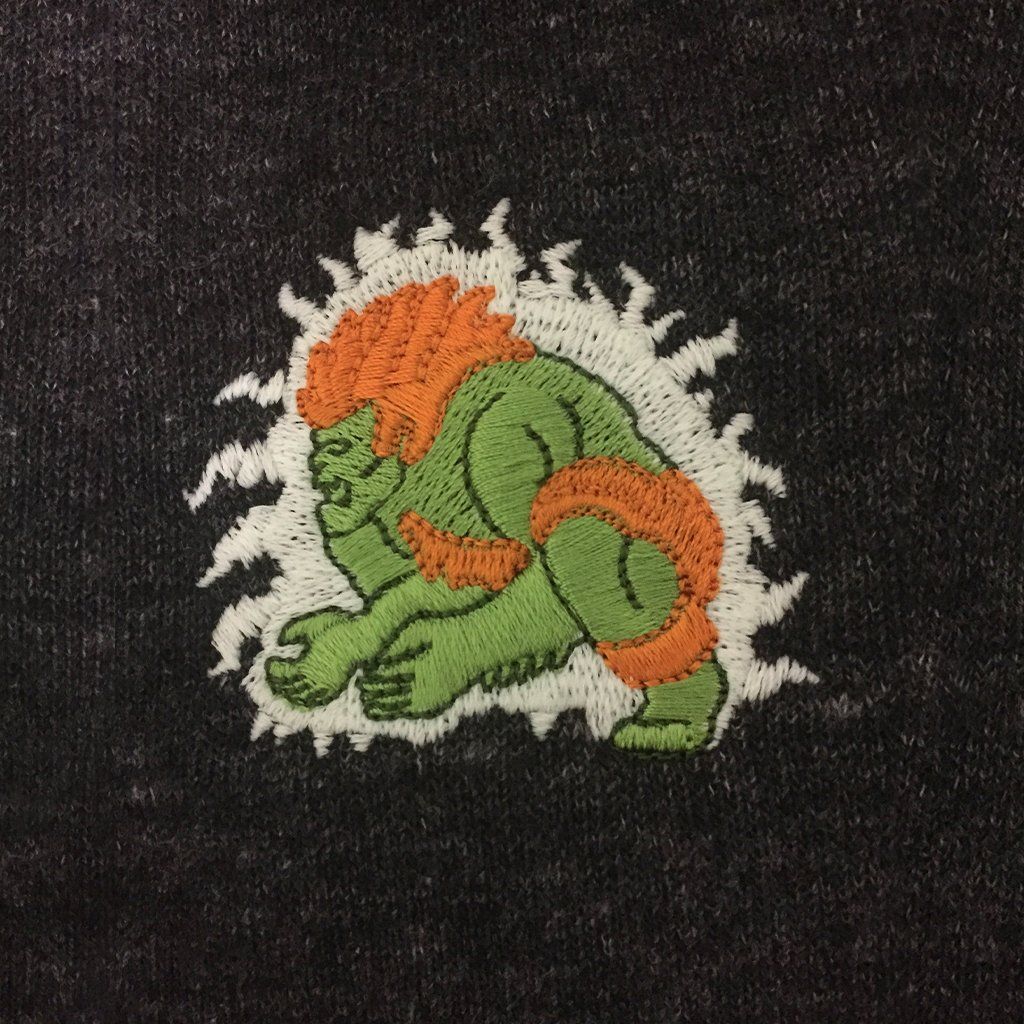 Close-up of green Blanka embroidery with orange mane on dark fabric.